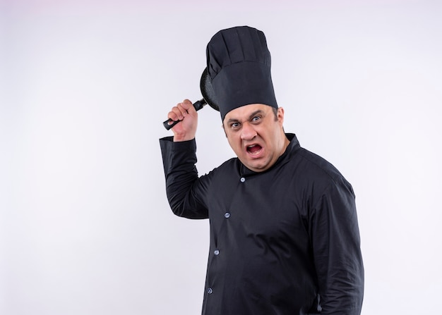 怒っている男性シェフは、黒い制服を着て、白い背景の上に立っている攻撃的な表情で鍋を振る帽子を調理します