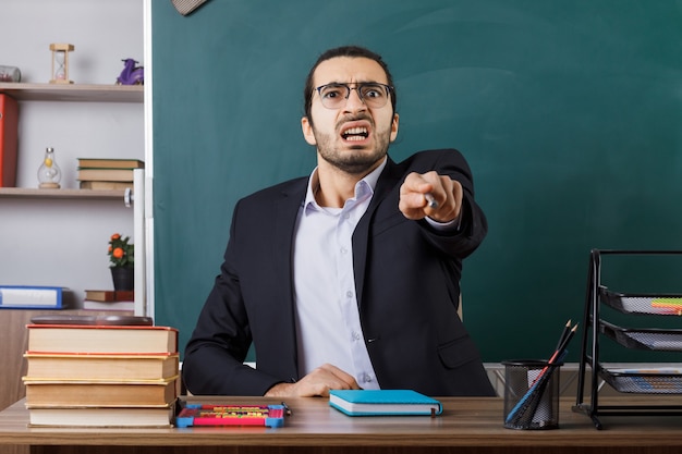 Angry male teacher wearing glasses points with pointer stick sitting at table with school tools in classroom