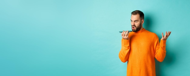 Angry man arguing on speakerphone record voice message with mad face standing over light blue backgr