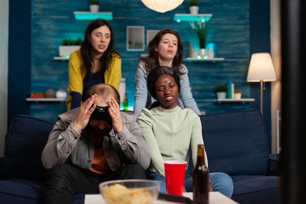 Angry man and multi ethnic friends upset after losing game competition, bonding and sitting on couch after drinking beer