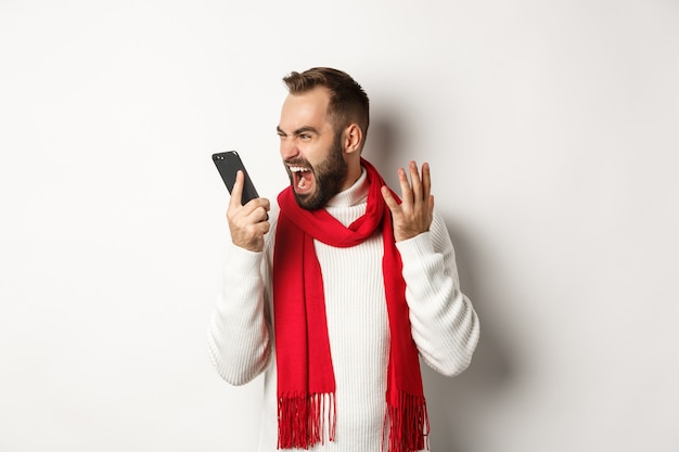 Angry man shouting at smartphone with mad face, standing furious against white background