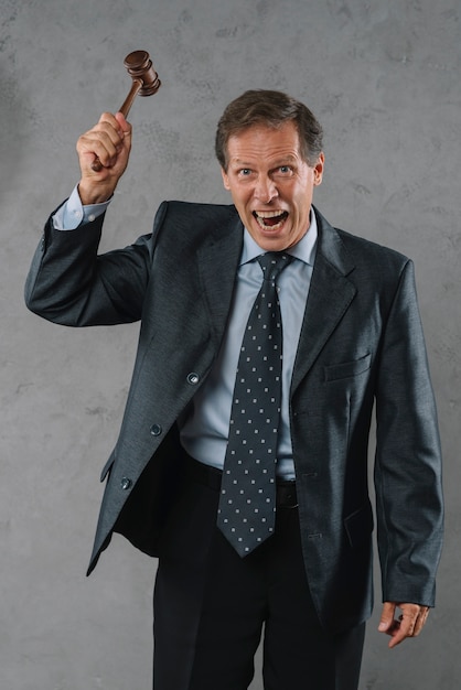 An angry mature male lawyer hitting with gavel against gray textured background