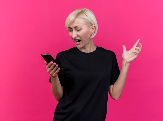 Angry middle-aged blonde woman holding and looking at mobile phone keeping hand in air isolated on pink wall