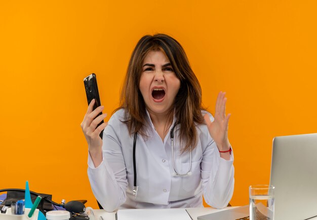 Angry middle-aged female doctor wearing medical robe and stethoscope sitting at desk with medical tools clipboard and laptop holding mobile phone keeping hand in air isolated