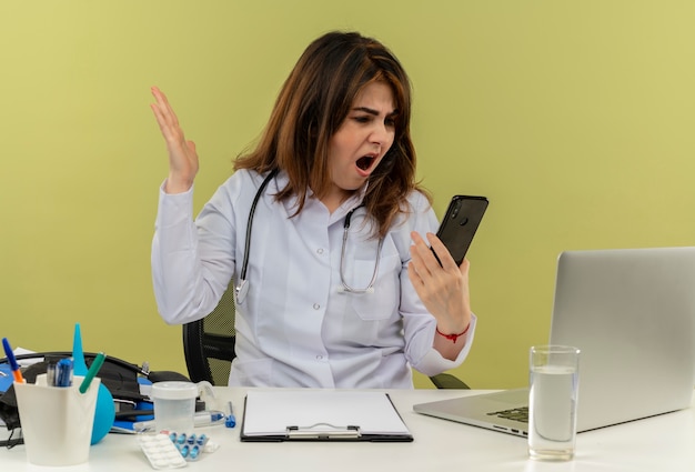 Angry middle-aged female doctor wearing medical robe with stethoscope sitting at desk work on laptop with medical tools holding and looking at phone on isolated green wall with copy space