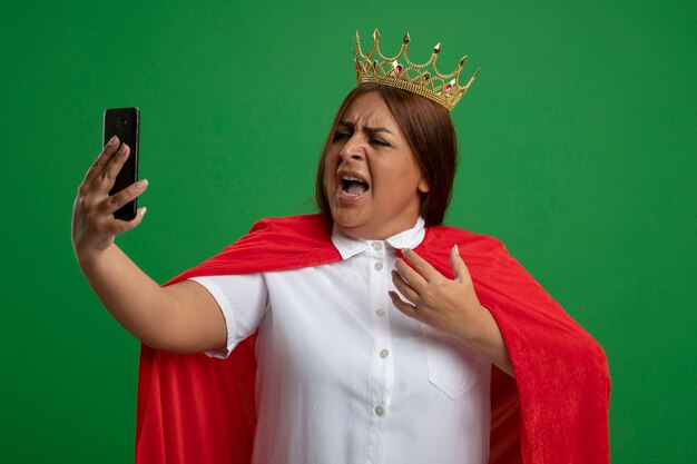 Angry middle-aged superhero female wearing crown holding and looking at phone isolated on green background
