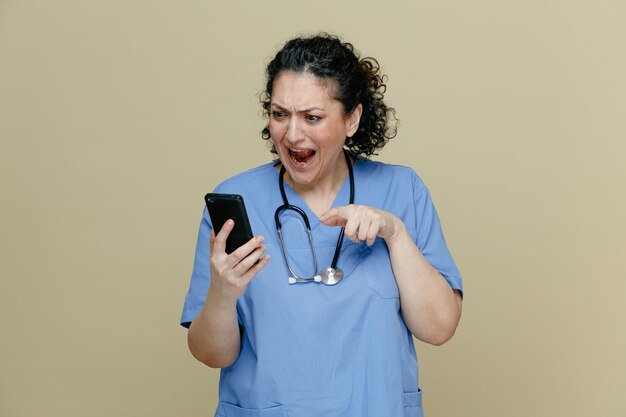 Angry middleaged female doctor wearing uniform and stethoscope around neck holding mobile phone looking at it and pointing at it shouting isolated on olive background