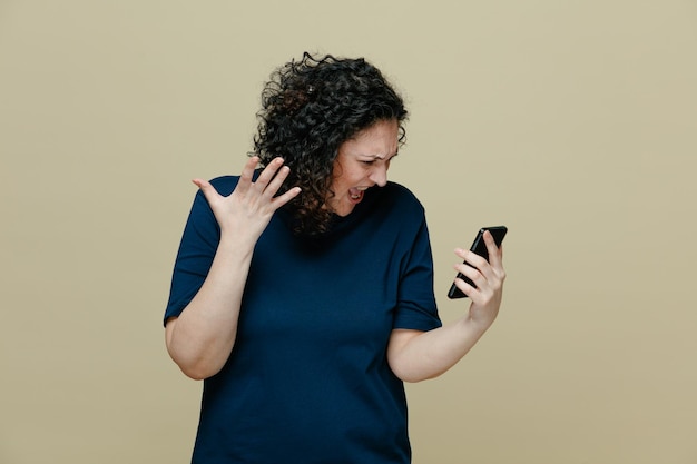 Foto gratuita donna arrabbiata di mezza età che indossa una maglietta che tiene il telefono cellulare guardandolo mostrando la mano vuota che grida isolato su sfondo verde oliva
