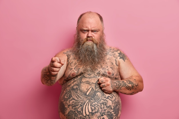 Angry offended overweight man keeps hand on big naked tummy and blames you, hears offensive words about himself, has tattooed body and long thick beard, made keep to diet