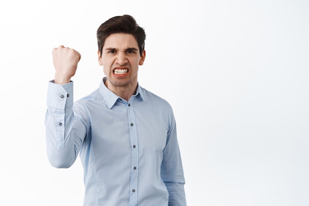 Сердитый офисный работник грозит кулаком, выглядит возмущенным и рассерженным, разочарованно стоит на белом фоне
