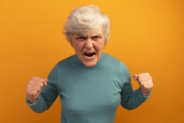 Сердитая старуха в синем свитере с высоким воротом, сжимая кулаки, кричит