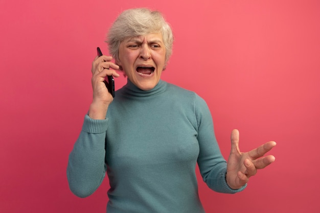 Angry old woman wearing blue turtleneck sweater talking on phone keeping hand in air looking down 