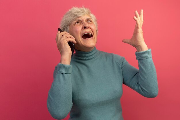 Angry old woman wearing blue turtleneck sweater talking on phone looking up raising hand up isolated on pink wall