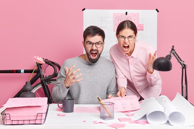 Angry outraged professional woman and man colleagues scream loudly being annoyed with difficult task pose at desktop surrounded by blueprints papers sketches collaborate for common building project