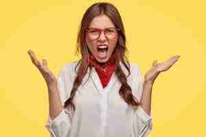 Free photo angry outraged woman shouts with spread hands, feels betrayed and pissed, expresses bad emotions, frowns face in annoyance, dressed in stylish clothing, isolated over yello wall, has conflict