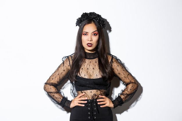 Free photo angry and pissed-off asian female magician, evil witch in black dress and wreath looking mad at someone, frowning and looking judgemental, standing over white background.