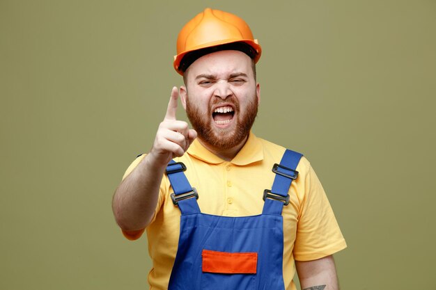 Angry points at camera young builder man in uniform isolated on green background