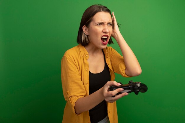 Angry pretty caucasian woman puts hand on head and holds controller looking at side on green