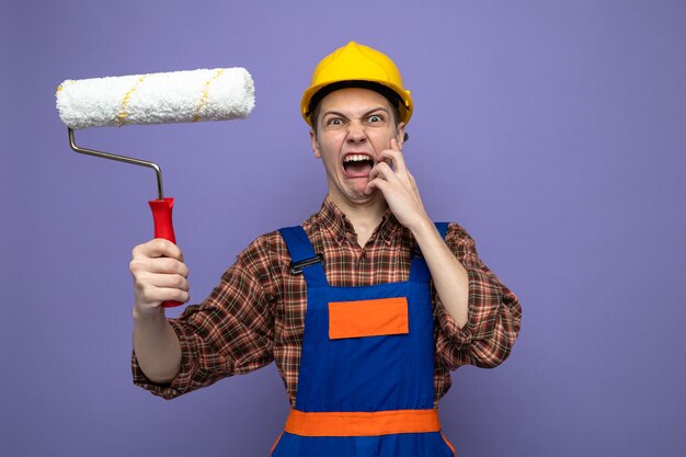 Angry putting hand on cheek young male builder wearing uniform holding roller brush 