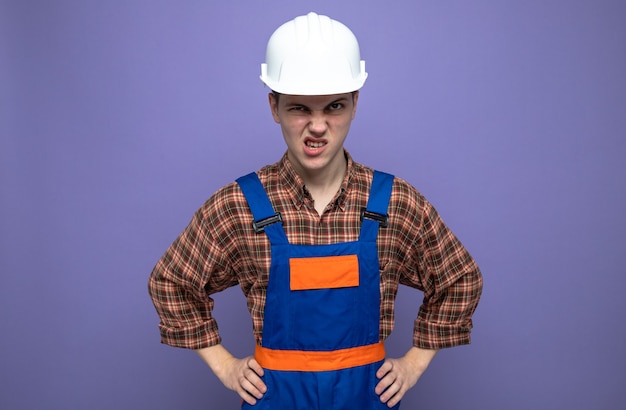 Angry putting hands on hip young male builder wearing uniform 