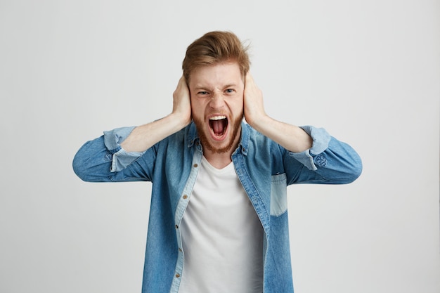 Angry rage young man with beard shouting screaming closing ears.