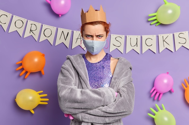 Free photo angry redhead european woman looks annoyed keeps arms folded wears disposable mask to protect from coronavirus dressed in domestic robe poses against purple wall with colorful balloons