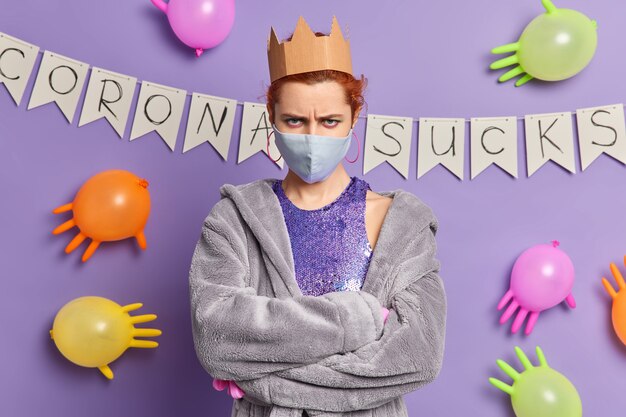 Angry redhead European woman looks annoyed keeps arms folded wears disposable mask to protect from coronavirus dressed in domestic robe poses against purple wall with colorful balloons