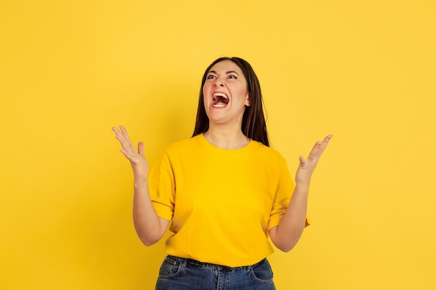 Angry screaming. Caucasian woman's portrait isolated on yellow  wall. Beautiful female brunette model in casual style. Concept of human emotions, facial expression, sales, ad, copyspace.