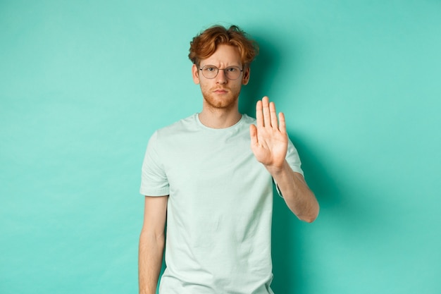 Angry and serious young man with red hair, wearing glasses, showing stop gesture, telling no, disapprove and prohibit something bad, standing over turquoise background.