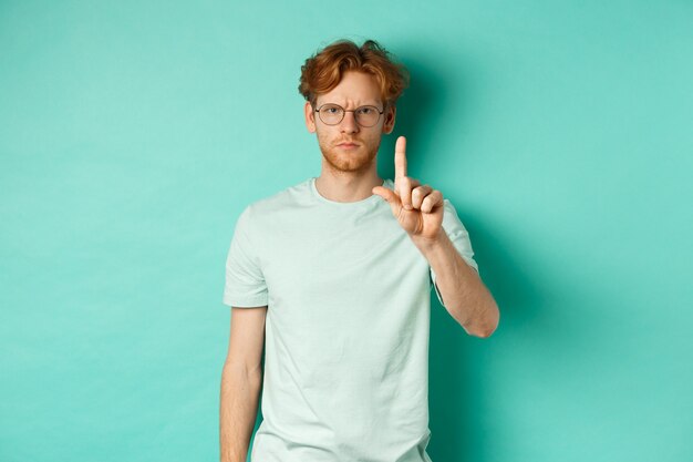 Сердитый и серьезный молодой человек с рыжими волосами, в очках, показывающий жест стоп, говорящий «нет», неодобрительно трясущий пальцем, стоящий на мятном фоне