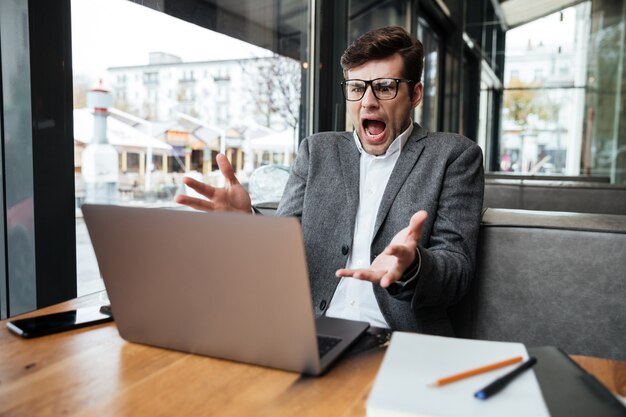 カフェでテーブルのそばに座ってラップトップコンピューターを見ながら眼鏡の怒っているショックを受けたビジネスマン