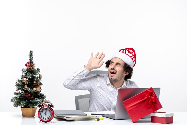 Angry shocked young businessman with funny santa claus hat looking above in the office on white background