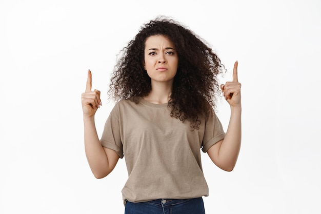 Angry silly brunette girl complains, points fingers up and sulking upset, staring disappointed or offended, displeased by advertisement, standing over white background.