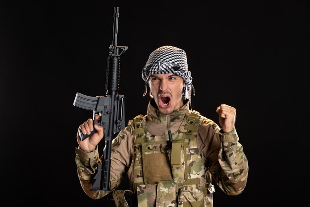 Angry soldier in camouflage holding machine gun on black wall