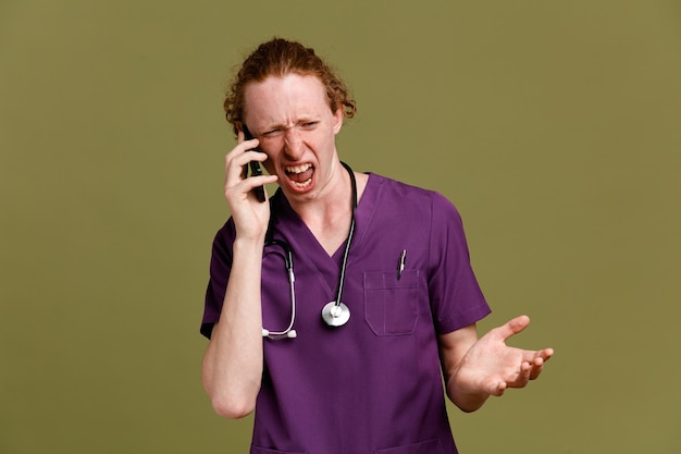 怒っている電話で話す若い男性医師は、緑の背景に分離された聴診器で制服を着ています