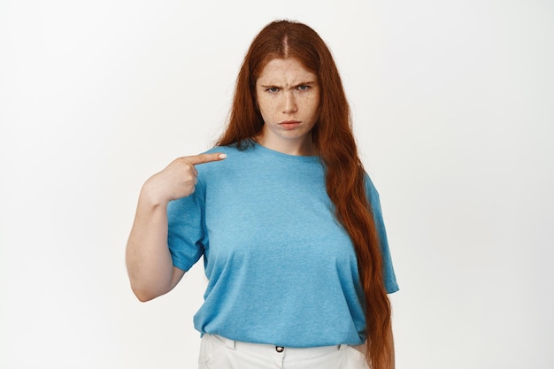 Angry sulking teen girl with red long hair, frowning upset, pointing finger at herself and complaining, staring displeased, standing against white background Premium Photo