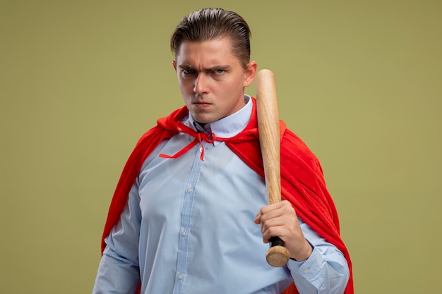 Angry super hero businessman in red cape holding baseball bat looking at camera with serious confident expression standing over light background
