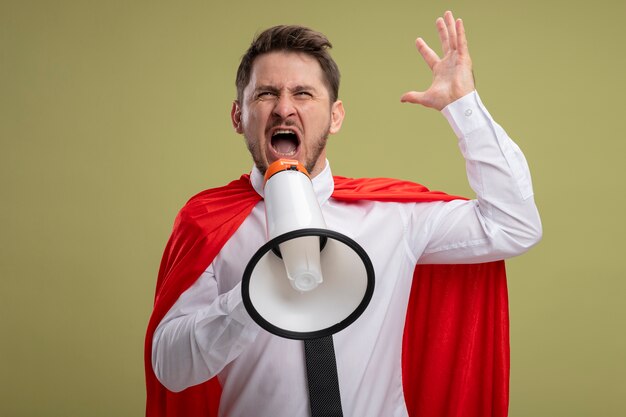 Angry super hero businessman in red cape shouting to megaphone with aggressive expression with raised arm standing over green background