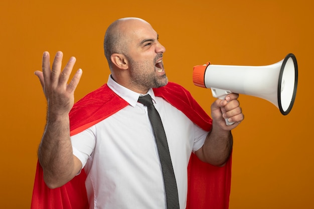 Uomo d'affari arrabbiato super eroe in mantello rosso che grida al megafono con il braccio alzato in piedi sopra la parete arancione