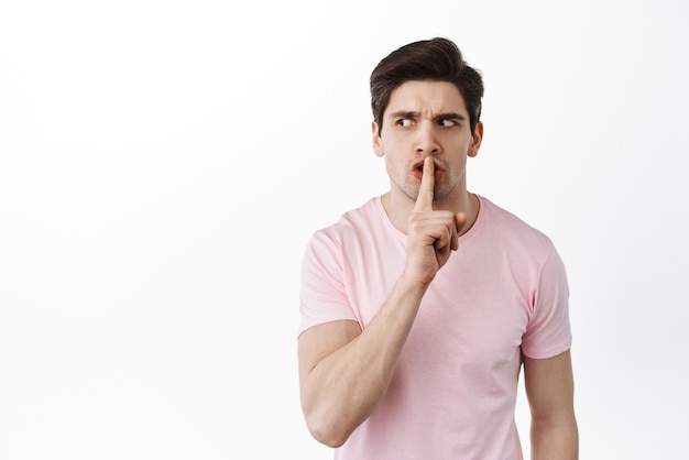 Angry suspicious man looks aside and shushing tell to shut up be quiet need silence show taboo gesture standing over white background