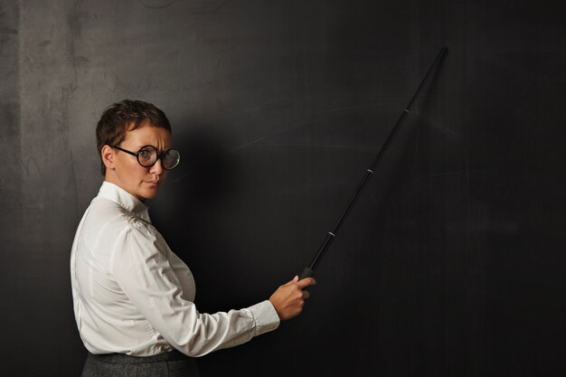 Angry teacher with pointer shows something on black chalk board