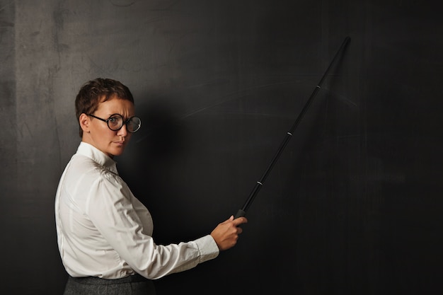 Angry teacher with pointer shows something on black chalk board