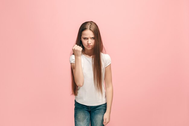 トレンディなピンクの壁に立っている怒っている十代の少女。女性の半身像。人間の感情、表情の概念。正面図。