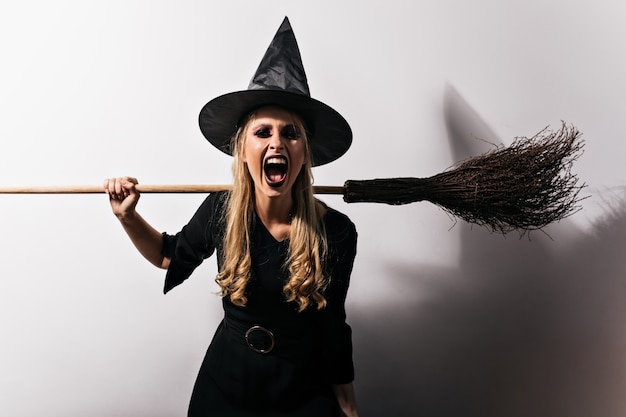 Free photo angry witch with long hair holding broom. blonde female wizard screaming in halloween.