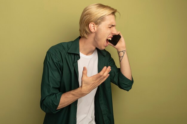 Злой с закрытыми глазами молодой блондин в зеленой футболке разговаривает по телефону