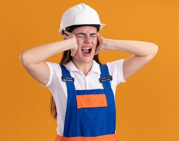 Free photo angry with closed eyes young builder woman in uniform putting hands on ears isolated on orange wall