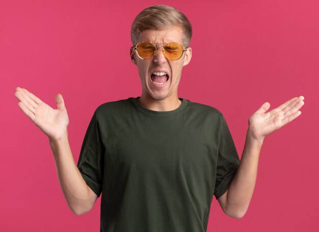Бесплатное фото Сердитый с закрытыми глазами молодой красивый парень в зеленой рубашке и очках, разводя руками, изолирован на розовой стене