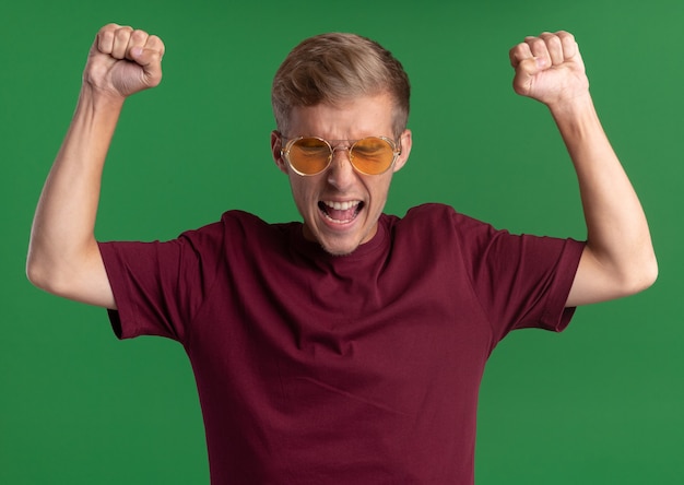 Foto gratuita arrabbiato con gli occhi chiusi giovane bel ragazzo che indossa la camicia rossa e occhiali alzando i pugni isolati sulla parete verde