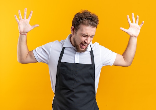 Free photo angry with closed eyes young male barber wearing uniform raising hands isolated on yellow wall