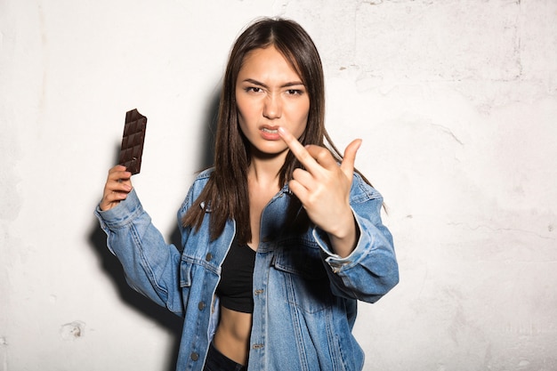 Сердитая женщина ест шоколад, показывая средний палец